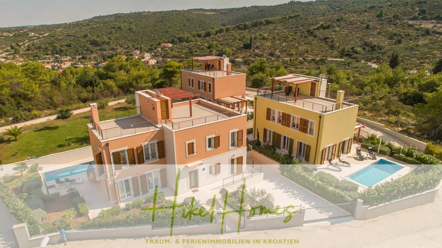 Villa im Mittelmeerstil mit 3 SZ, Pool, Dachterrasse mit Meerblick (3/4) - Luftaufnahme einer modernen Villa mit gelber und terrakottafarbener Fassade, mehreren Balkonen, einem eingezäunten Hof und einem Swimmingpool, umgeben von Grünflächen und Hügeln in Kroatien. Der Schriftzug "Haus Homes" ist am unteren Rand eingeblendet. (45739)