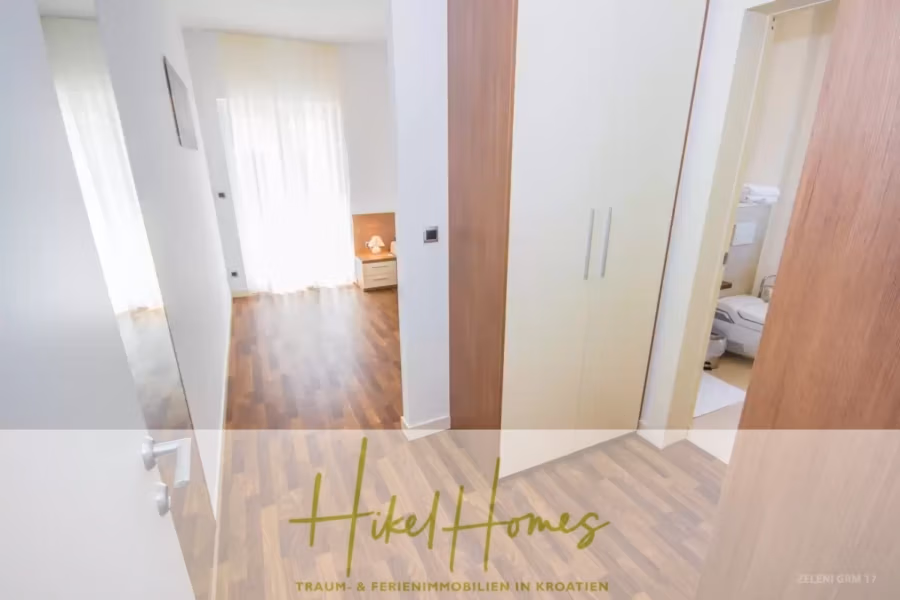 Villa im Mittelmeerstil mit 3 SZ, Pool, Dachterrasse mit Meerblick (3/4) - Ein heller Flur mit Holzboden führt links zu einem Schlafzimmer mit Bett und Tageslicht durch große Vorhänge. Rechts gibt es einen Blick auf ein Badezimmer mit Handtüchern. Unten steht „Hikel Homes“ und „Traum- & Ferienimmobilien in Kroatien“. (43317)