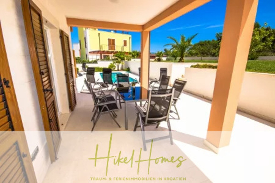 Villa im Mittelmeerstil mit 3 SZ, Pool, Dachterrasse mit Meerblick (3/4) - Eine überdachte Terrasse mit einem Tisch und Stühlen mit Blick auf einen privaten Pool und Garten. Modernes Haus und Palmen sind unter einem klaren blauen Himmel zu sehen. Der Text lautet "Hikel Homes - Traum- & Ferienimmobilien in Kroatien. (45746)