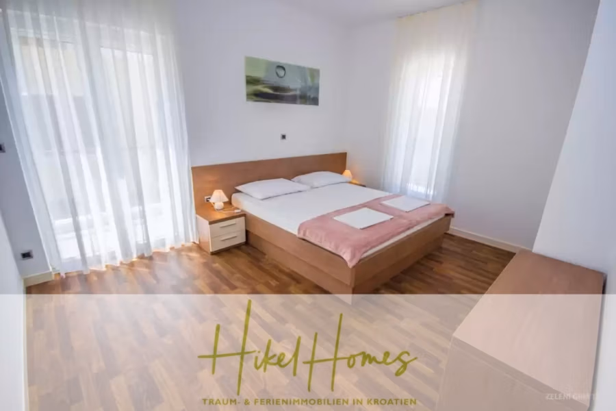 Villa im Mittelmeerstil mit 3 SZ, Pool, Dachterrasse mit Meerblick (3/4) - Minimalistisches Schlafzimmer mit einem Doppelbett mit weißer Bettwäsche und einer rosafarbenen Decke, flankiert von zwei Nachttischen. Sonnenlicht fällt durch transparente Vorhänge an großen Fenstern und erhellt den Holzboden. Über dem Bett hängt ein Landschaftsgemälde. (43314)