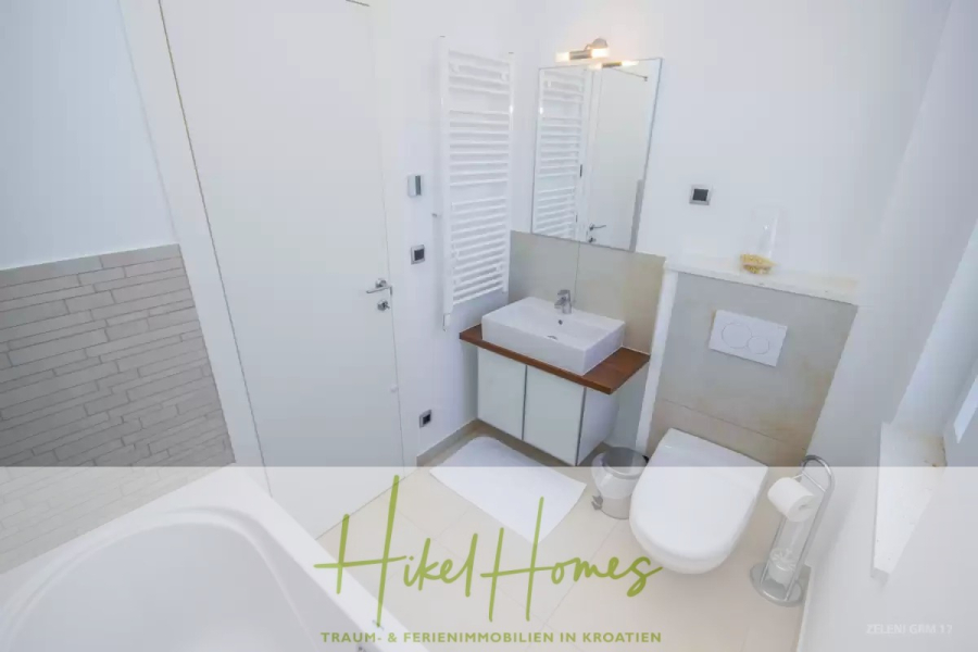 Villa im Mittelmeerstil mit 3 SZ, Pool, Dachterrasse mit Meerblick (3/4) - Modernes weißes Badezimmer mit Badewanne, Waschbecken, Spiegel, Wandtoilette, Handtuchheizkörper und kleinem Abfalleimer; Schriftzug "Hikel Homes" am Boden. (45754)