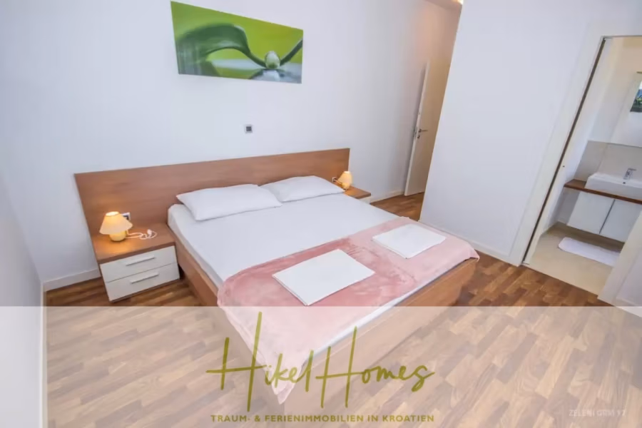 Villa im Mittelmeerstil mit 3 SZ, Pool, Dachterrasse mit Meerblick (3/4) - Modernes Schlafzimmer mit Doppelbett und weißer und rosa Bettwäsche. Das Zimmer verfügt über Holzboden, zwei Nachttische mit Lampen und ein grünes abstraktes Wandbild. Eine offene Tür führt zum Badezimmer mit ebenerdiger Dusche. (43321)