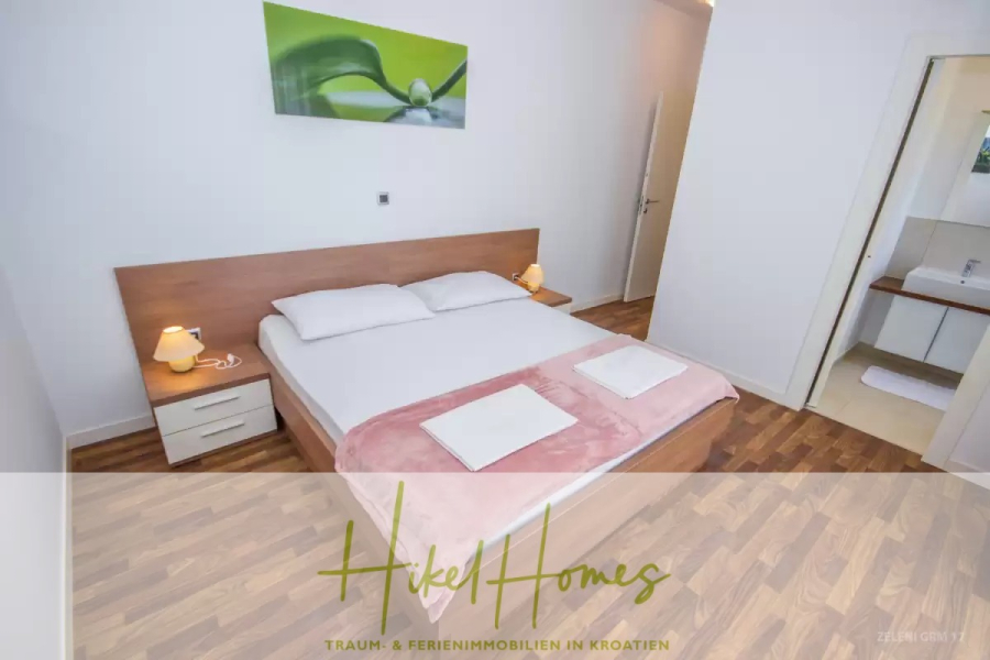 Villa im Mittelmeerstil mit 3 SZ, Pool, Dachterrasse mit Meerblick (3/4) - Ein modernes, minimalistisches Schlafzimmer mit einem Doppelbett, zwei Nachttischen mit Lampen, gefalteten Handtüchern auf einer rosafarbenen Decke, Holzfußboden und einer offenen Tür, die zu einem Badezimmer führt. Der Schriftzug "Hikel Homes" ist am unteren Rand eingeblendet. (45762)