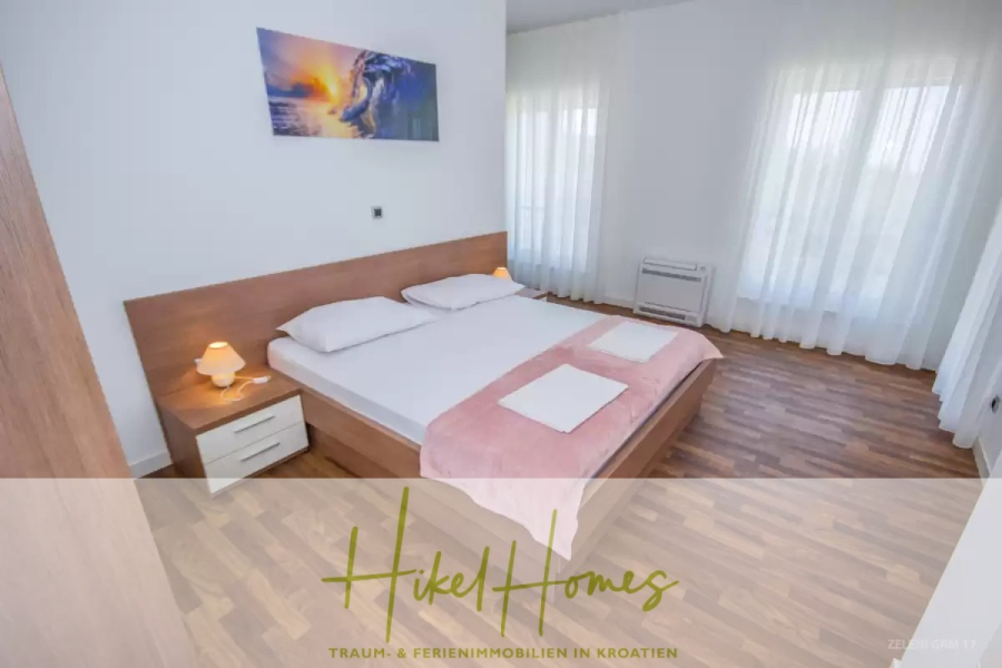 Villa im Mittelmeerstil mit 3 SZ, Pool, Dachterrasse mit Meerblick (3/4) - Ein helles Schlafzimmer mit einem Doppelbett, weißer Bettwäsche, rosa Überwurf und Handtüchern, Holzmöbeln, zwei Nachttischlampen und durchsichtigen Vorhängen vor großen Fenstern. Über dem Bett hängt ein buntes Kunstwerk. (45764)