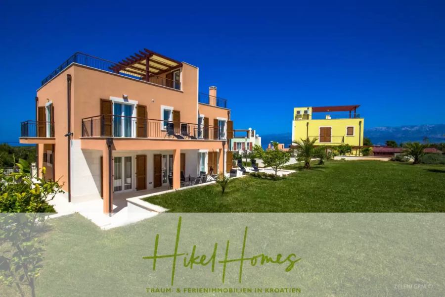 Villa im Mittelmeerstil mit 3 SZ, Pool, Dachterrasse mit Meerblick (3/4) - Zwei farbenfrohe moderne Häuser mit Balkonen und Terrassen stehen auf einem hellgrünen Rasen unter einem klaren blauen Himmel, mit Bergen im Hintergrund. Der überlagerte Text lautet "Hikel Homes: Traum- & Ferienimmobilien in Kroatien. (45743)