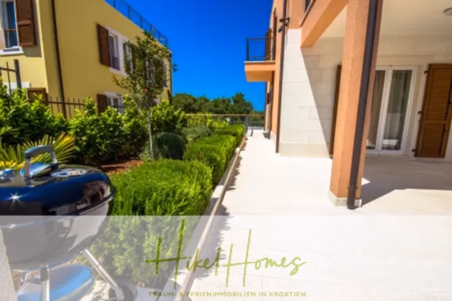 Villa im Mittelmeerstil mit 3 SZ, Pool, Dachterrasse mit Meerblick (3/4) - Eine sonnendurchflutete Terrasse neben einem modernen Gebäude mit orange-weißen Wänden, einem glänzenden silbernen Grill, üppigen grünen Sträuchern und einem kleinen Rasen. Links ist ein Nachbargebäude mit Holzläden zu sehen. Unten ist ein Logo mit Text zu sehen. (43307)