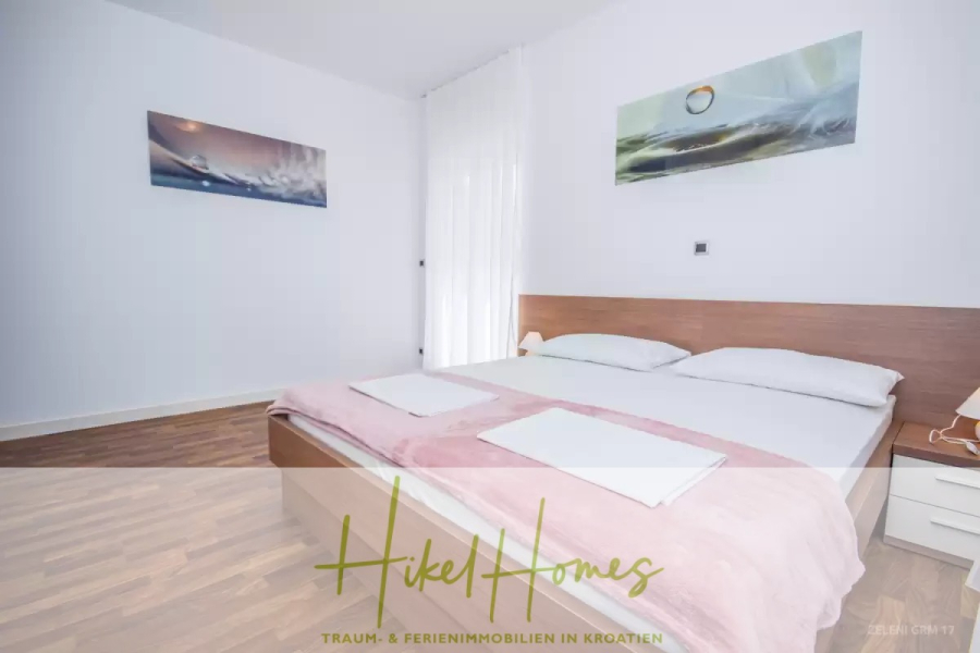 Villa im Mittelmeerstil mit 3 SZ, Pool, Dachterrasse mit Meerblick (3/4) - Ein helles, minimalistisches Schlafzimmer mit einem Doppelbett, einer hellrosa Decke, zwei Handtüchern und weißen Wänden. Es gibt zwei abstrakte Gemälde, ein Fenster mit durchsichtigen Vorhängen und einen Holzfußboden. Die Textüberlagerung lautet "Hikel Homes". (45756)