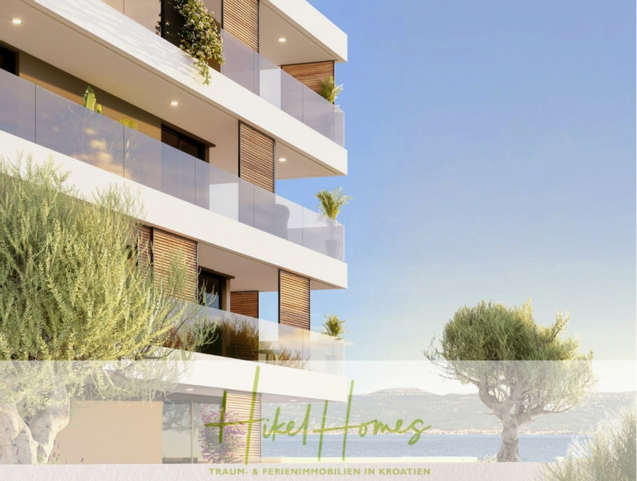 1. Reihe hochwertige Neubau-Meerblick Wohnung mit 1 SZ, Terrasse und Gemeinschaftspool (S2B) - Modernes Apartmentgebäude am Strand mit Glasbalkonen, Topfpflanzen und Bäumen, mit Blick auf das Meer. Das Logo "Hikel Homes" und deutscher Text erscheinen am unteren Rand des Bildes. (46164)