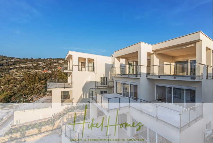 5 Sterne Villa Bruna – Meerblick auf 260m² WFL in 1. Reihe / 4 SZ / Neubau / Sofort bezugsfertig - Moderne weiße Wohnhäuser mit Glasbalkonen vor einer malerischen Kulisse aus Hügeln und Bäumen unter einem klaren blauen Himmel. Unten steht das Logo „Hikel Homes Traum- & Ferienimmobilien in Kroatien“. (41405)
