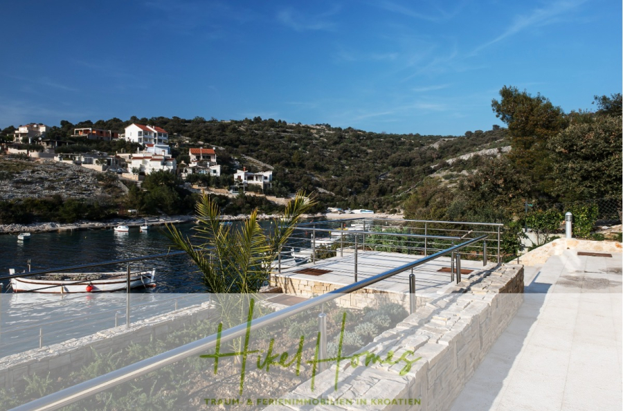 5 Sterne Villa Bruna – Meerblick auf 260m² WFL in 1. Reihe / 4 SZ / Neubau / Sofort bezugsfertig - Blick auf eine ruhige Uferpromenade mit angedockten Booten, Häusern am Hang im Hintergrund und einer Steinterrasse mit Metallgeländer im Vordergrund unter einem klaren blauen Himmel. (46371)