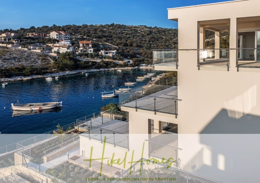 5 Sterne Villa Bruna – Meerblick auf 260m² WFL in 1. Reihe / 4 SZ / Neubau / Sofort bezugsfertig - Ein modernes Küstengebäude mit Glasbalkonen überblickt eine ruhige Bucht mit mehreren kleinen Booten. Auf der anderen Seite des Wassers liegen von Grün umgebene Häuser am Hang. Unten im Bild ist das Logo eines Immobilienunternehmens zu sehen. (41407)