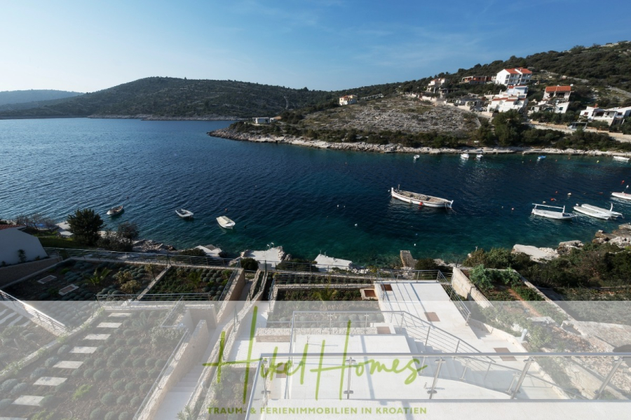 5 Sterne Villa Bruna – Meerblick auf 260m² WFL in 1. Reihe / 4 SZ / Neubau / Sofort bezugsfertig - Blick auf eine ruhige blaue Bucht mit Booten, felsigen Hügeln und Häusern entlang des Ufers in Kroatien, gesehen von einer Terrasse mit Blick auf einen Garten. Am unteren Bildrand sind "Hotel Homes" und deutscher Text zu sehen. (46366)