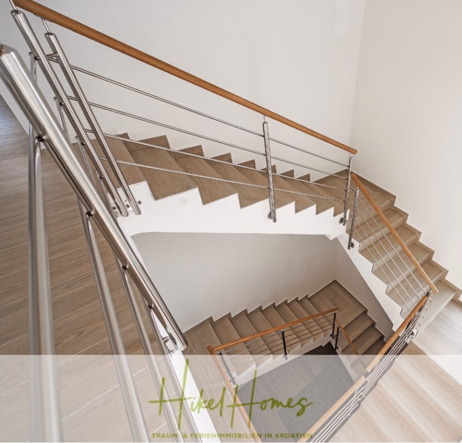 5 Sterne Villa Bruna – Meerblick auf 260m² WFL in 1. Reihe / 4 SZ / Neubau / Sofort bezugsfertig - Eine moderne Holztreppe mit Metallgeländer führt von oben betrachtet spiralförmig nach unten. Der Raum ist minimalistisch gestaltet und in natürlichen Holztönen gehalten. Logo und Text unten zeigen „Vikel Homes“ mit einem Slogan über Ferienimmobilien in Kroatien. (41417)