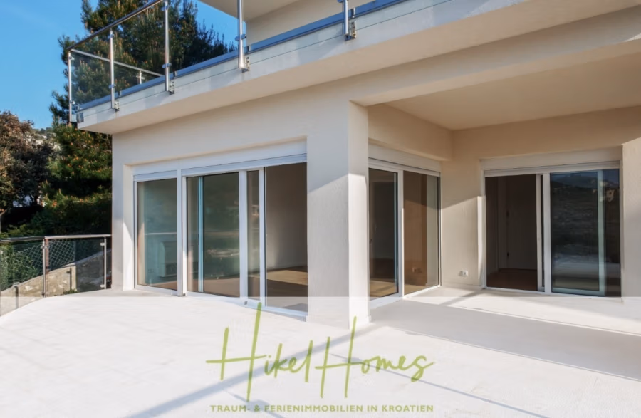 5 Sterne Villa Bruna – Meerblick auf 260m² WFL in 1. Reihe / 4 SZ / Neubau / Sofort bezugsfertig - Ein modernes Haus mit großen Glasschiebetüren, die auf eine geräumige weiße Terrasse führen. Das Gebäude hat eine hellbeige Fassade und einen Glasbalkon im Obergeschoss. Im Hintergrund sind Bäume zu sehen. (41402)