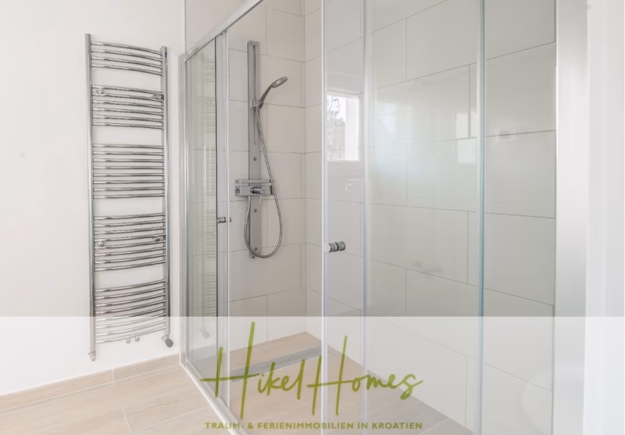 5 Sterne Villa Bruna – Meerblick auf 260m² WFL in 1. Reihe / 4 SZ / Neubau / Sofort bezugsfertig - Minimalistisches Badezimmer mit verglaster Dusche und modernem Duschkopf. Links ein vertikaler Handtuchwärmer. Helle Fliesen bedecken Boden und Wände. Der Markentext unten lautet „Hikel Homes – Traum & Ferienimmobilien in Kroatien“. (41419)