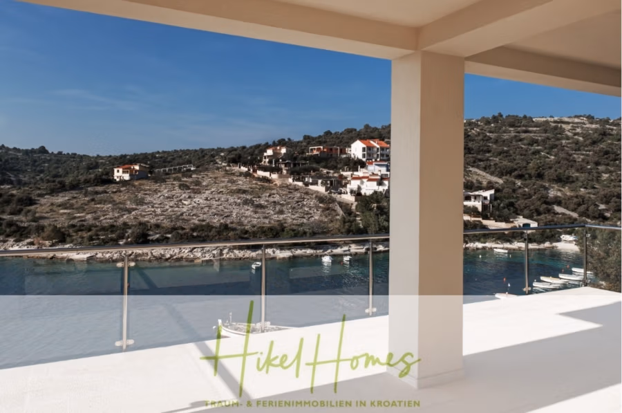 5 Sterne Villa Bruna – Meerblick auf 260m² WFL in 1. Reihe / 4 SZ / Neubau / Sofort bezugsfertig - Blick von einem Balkon auf eine ruhige, blaue Uferpromenade mit einem mit Häusern übersäten Hügel. Der Himmel ist klar, die Szenerie friedlich. Ein transparentes Geländer säumt den Balkon. Unten befinden sich ein Logo und ein Text, die die Szene teilweise verdecken. (41401)