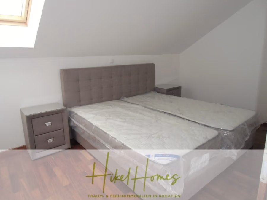 Appartement über 2 Stockwerke / 3 Schlafzimmer / 70m zum Meer / Pool / große Terrasse - Ein modernes Schlafzimmer mit schräger Decke verfügt über ein Doppelbett mit gepolstertem Kopfteil. Das Bett hat kunststoffbezogene Matratzen und wird von zwei weißen Nachttischen flankiert. Ein Oberlicht sorgt für Tageslicht. Unten ist das Logo „Hikel Homes“ eingelegt. (41231)