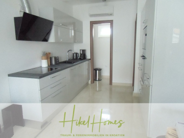 Moderne, minimalistische Küche mit weißen Schränken, schwarzen Arbeitsplatten, Einbaugeräten und kleinen Küchengeräten auf der Theke. Ein Fenster und ein Mülleimer befinden sich am anderen Ende. Der Schriftzug "Hikel Homes" ist am unteren Rand eingeblendet. - Appartement über 2 Stockwerke / 3 Schlafzimmer / 70m zum Meer / Pool / große Terrasse