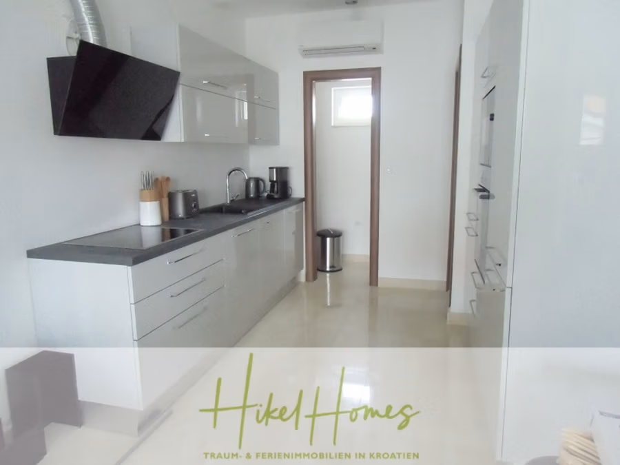Appartement über 2 Stockwerke / 3 Schlafzimmer / 70m zum Meer / Pool / große Terrasse - Moderne, elegante Küche mit glänzend weißen Schränken, dunklen Arbeitsplatten und Edelstahlgeräten, darunter Backofen und Kaffeemaschine. Über dem Herd befindet sich eine Dunstabzugshaube. Ein Fenster am Ende sorgt für Tageslicht. Der Hikel Homes-Schriftzug ist unten eingeblendet. (41226)