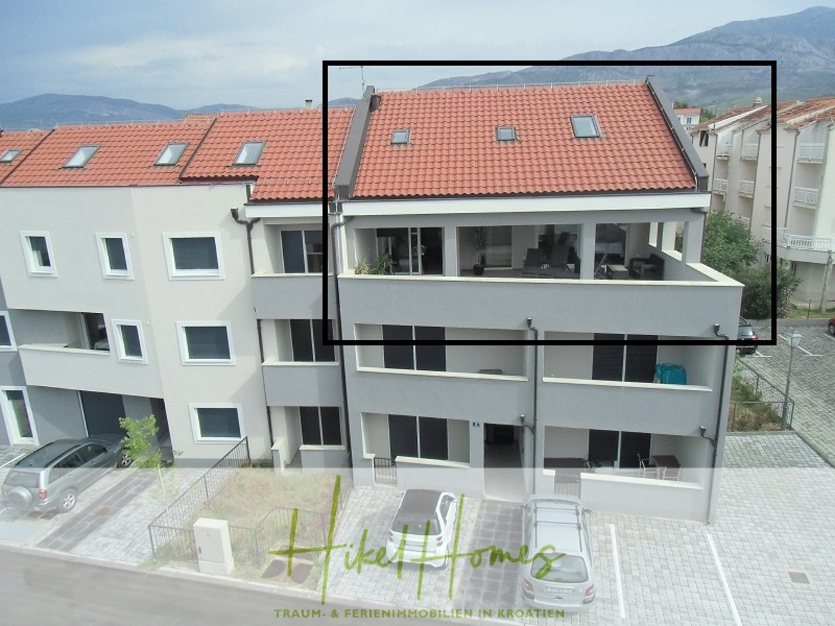 Ein modernes Mehrfamilienhaus mit rotem Ziegeldach, grauer und weißer Fassade und Balkonen. Eine Wohnung im Obergeschoss mit einer großen Terrasse ist schwarz umrandet. Im Hintergrund sind Berge und andere Gebäude zu sehen. - Appartement über 2 Stockwerke / 3 Schlafzimmer / 70m zum Meer / Pool / große Terrasse