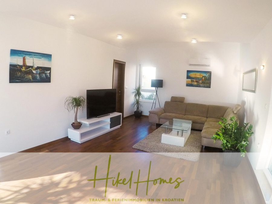 Appartement über 2 Stockwerke / 3 Schlafzimmer / 70m zum Meer / Pool / große Terrasse - Modernes Wohnzimmer mit einem Sektionssofa, einem Fernseher auf einem weißen Ständer, einem Couchtisch, Wandkunst, einer Topfpflanze und Holzfußboden. Sonnenlicht strömt herein und schafft einen hellen, einladenden Raum. Der Schriftzug "Hikel Homes" befindet sich am unteren Rand. (46632)