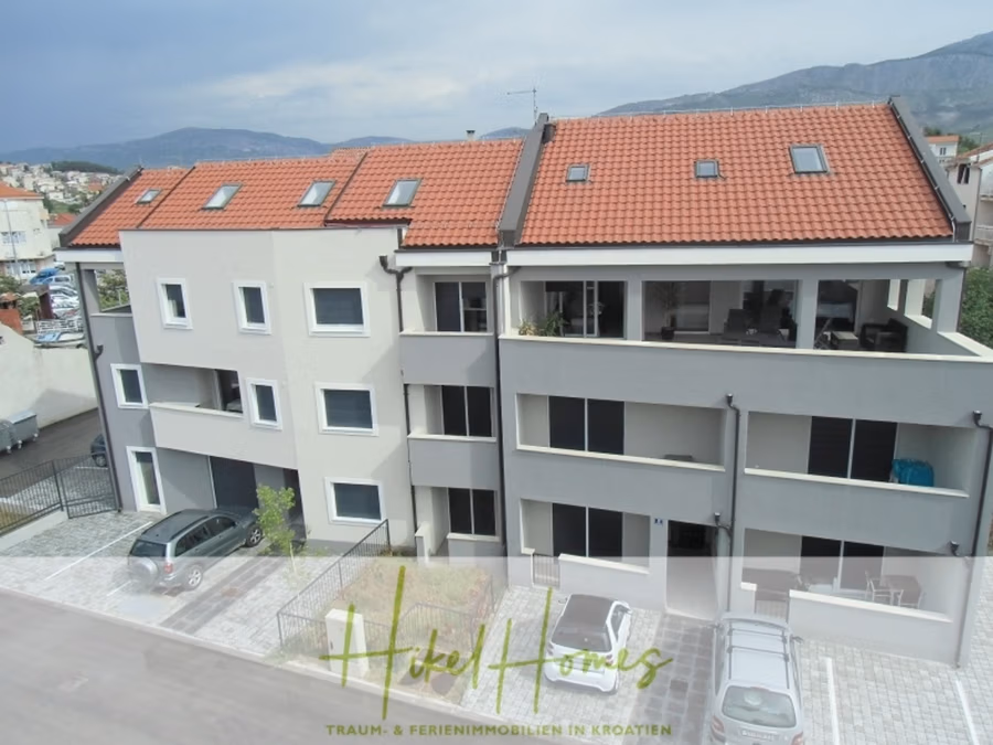 Appartement über 2 Stockwerke / 3 Schlafzimmer / 70m zum Meer / Pool / große Terrasse - Ein modernes, dreistöckiges Wohnhaus mit rotem Ziegeldach und großem Balkon. Jede Etage verfügt über rechteckige Fenster. Das Gebäude liegt vor einer Hügelkulisse und einem teilweise bewölkten Himmel. Im Vordergrund befindet sich ein gepflasterter Parkplatz. (41225)