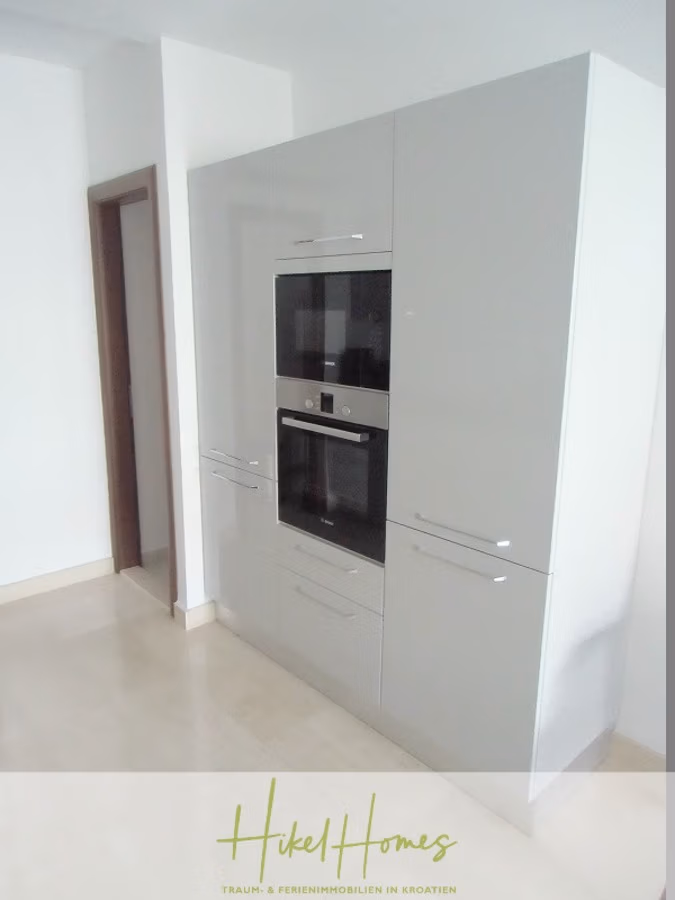 Appartement über 2 Stockwerke / 3 Schlafzimmer / 70m zum Meer / Pool / große Terrasse - Moderne Küche mit eingebautem Edelstahlbackofen und Mikrowelle. Die schlichten, weißen Schränke heben sich vom hellen Boden ab. Links ist eine Holztür zu sehen. Unten steht: „Hikel Homes – Traum- & Ferienimmobilien in Kroatien.“. (41227)