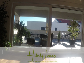 Ein moderner Balkon mit Terrassenmöbeln und Topfpflanzen durch große Glastüren im Sonnenlicht gesehen. Das Logo am unteren Rand lautet "Hikel Homes - Traum & Ferienimmobilien in Kroatien". - Appartement über 2 Stockwerke / 3 Schlafzimmer / 70m zum Meer / Pool / große Terrasse