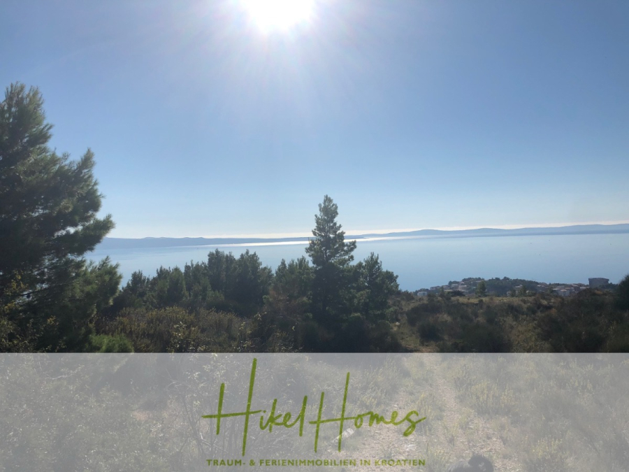 511m² Traumvilla auf über 3100m² großem unverbaubaren Meerblick Grundstück – eigene Auffahrt - Blick auf eine sonnige Küstenlandschaft mit grünen Bäumen und Sträuchern im Vordergrund, einem ruhigen blauen Meer und fernen Bergen unter einem klaren Himmel. Der Text am unteren Rand lautet: "Hikel Homes. TRAUM- & FERIENIMMOBILIEN IN KROATIEN.". (46428)