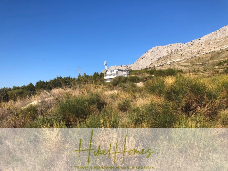 511m² Traumvilla auf über 3100m² großem unverbaubaren Meerblick Grundstück – eigene Auffahrt - Ein einsames Haus liegt eingebettet in einer Graslandschaft mit einem felsigen Hügel im Hintergrund unter einem klaren blauen Himmel. Grüne und gelbe Vegetation umgibt das Gebiet. Am unteren Bildrand ist der Text „Hikel Homes“ zu sehen. (41953)