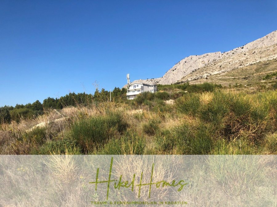 511m² Traumvilla auf über 3100m² großem unverbaubaren Meerblick Grundstück – eigene Auffahrt - Ein weißes Haus steht allein auf einem grasbewachsenen Hügel mit felsigen Bergen im Hintergrund unter einem klaren blauen Himmel. Im Vordergrund befinden sich hohe grüne und gelbliche Büsche. Die Worte "Hikel Homes" erscheinen am unteren Rand des Bildes. (46438)