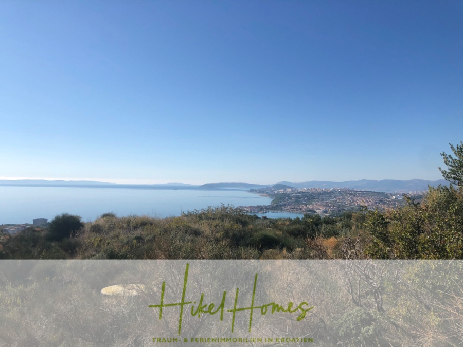 511m² Traumvilla auf über 3100m² großem unverbaubaren Meerblick Grundstück – eigene Auffahrt - Blick von einer Hügelkuppe über eine Küstenstadt mit blauem Himmel, ruhigem Meer und fernen Hügeln. Im Vordergrund ist die Vegetation zu sehen, und ein blasses Overlay am unteren Rand zeigt den Text "Hikel Homes". (46426)