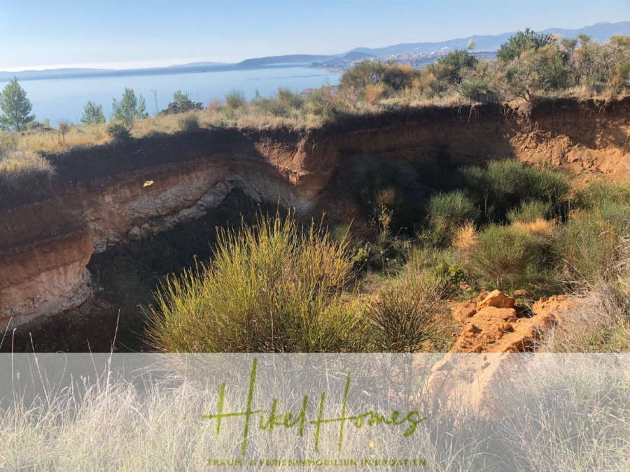 511m² Traumvilla auf über 3100m² großem unverbaubaren Meerblick Grundstück – eigene Auffahrt - Eine große Senke, umgeben von trockenem Gras und Sträuchern, liegt am Rande eines ruhigen blauen Sees, mit fernen Bergen und einem klaren Himmel im Hintergrund. Auf dem Boden steht "Hikel Homes". (46434)