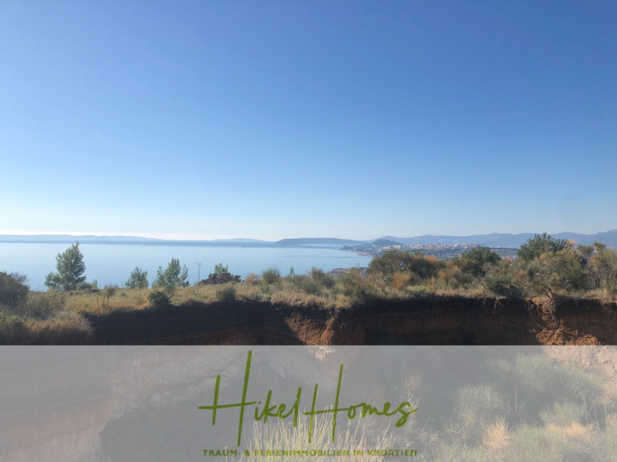 511m² Traumvilla auf über 3100m² großem unverbaubaren Meerblick Grundstück – eigene Auffahrt - Eine malerische Küstenlandschaft in Kroatien mit Sträuchern und Bäumen im Vordergrund, ruhigem blauen Meer und fernen Hügeln unter einem klaren Himmel. Der Text "Hikel Homes" erscheint am unteren Rand. (46432)
