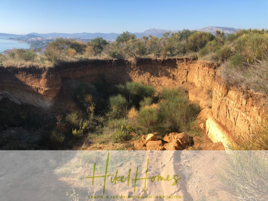 511m² Traumvilla auf über 3100m² großem unverbaubaren Meerblick Grundstück – eigene Auffahrt - Eine große, sonnenbeschienene Erdgrube mit steilen, exponierten Seiten und spärlicher Vegetation, umgeben von Sträuchern und Hügeln. In der Ferne sind Berge und ein Gewässer unter einem klaren blauen Himmel zu erkennen. (46435)