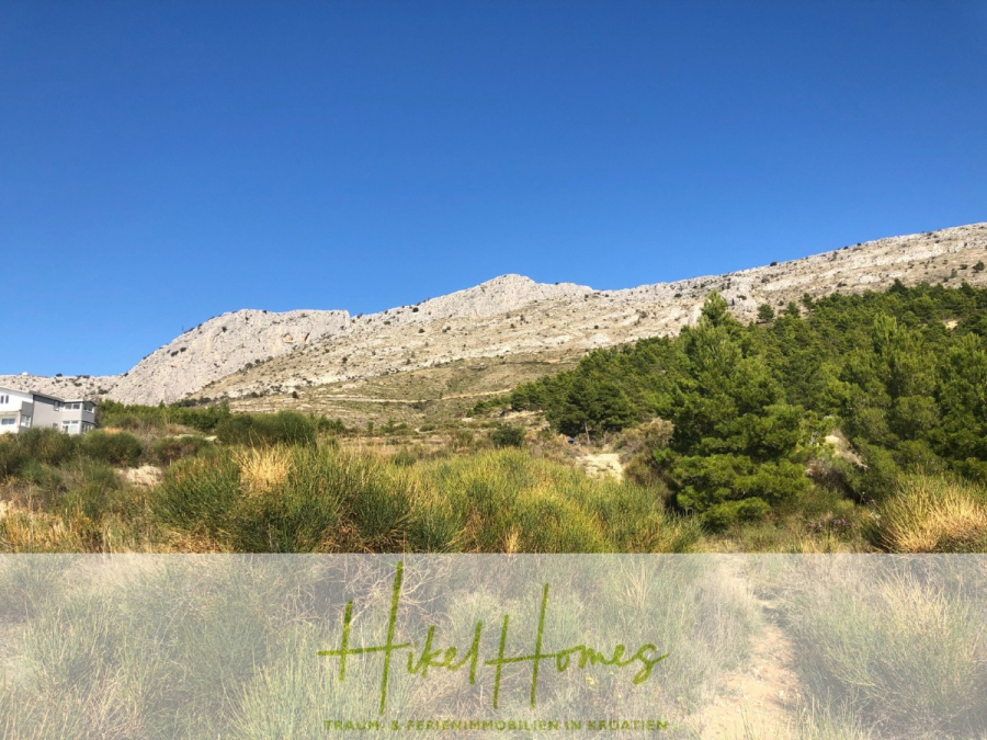 511m² Traumvilla auf über 3100m² großem unverbaubaren Meerblick Grundstück – eigene Auffahrt - Ein felsiger Berg unter einem klaren blauen Himmel, mit grünen Büschen und Bäumen im Vordergrund. Der Text am unteren Rand lautet "Hikel Homes - Traumhafte Ferienimmobilien in Kroatien". (46439)