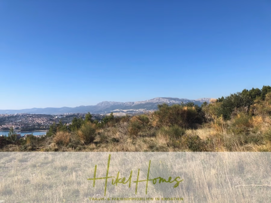 511m² Traumvilla auf über 3100m² großem unverbaubaren Meerblick Grundstück – eigene Auffahrt - Eine malerische Landschaft mit Grasfeldern im Vordergrund und einem Fernblick auf eine Stadt unter klarem blauen Himmel. Im Hintergrund sind Hügel oder Berge zu sehen. Unten befindet sich ein Wasserzeichen mit der Aufschrift „Hike Homes“. (41955)
