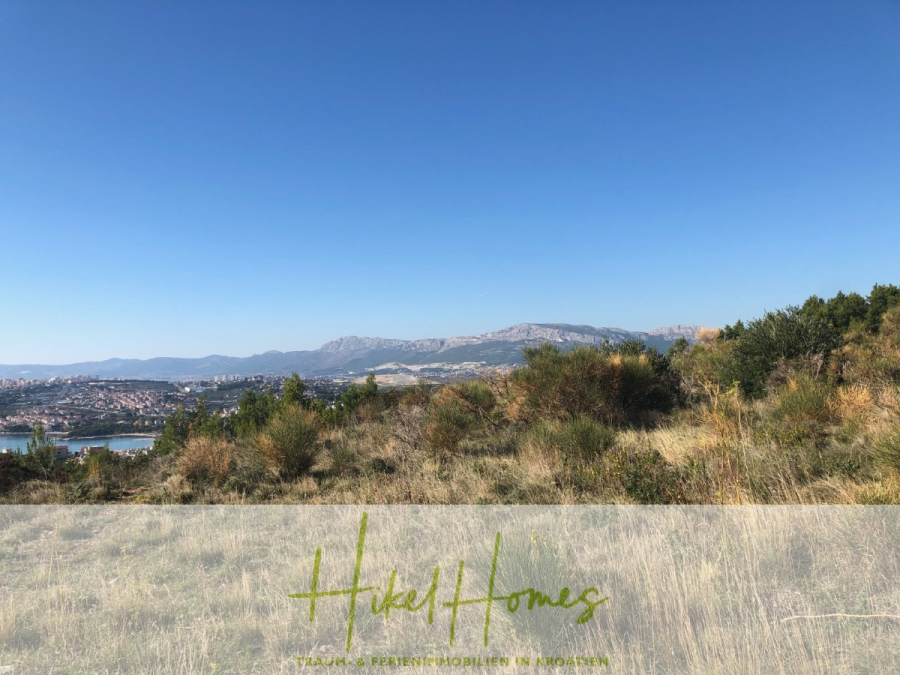 511m² Traumvilla auf über 3100m² großem unverbaubaren Meerblick Grundstück – eigene Auffahrt - Eine grasbewachsene, hügelige Landschaft unter einem klaren, blauen Himmel mit fernen Bergen und einer Stadt in Sichtweite; unten ist der Text "Hikel Homes, Travel & Present Mobilities in Modernity" eingeblendet. (46440)