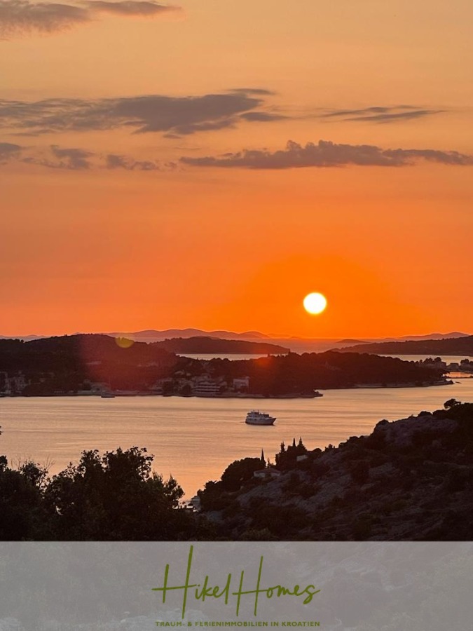 Prämiertes Architekturprojekt – Meerblick Luxus auf 490m² auf 1800m² Grund mit 2 Pools, Bar, Lounge - Ein leuchtend oranger Sonnenuntergang über einer ruhigen Bucht in Kroatien, mit Booten auf dem Wasser und Inseln, die sich vom Himmel abheben. Das "Hikel Homes"-Logo und der deutsche Text sind am unteren Rand des Bildes zu sehen. (45075)