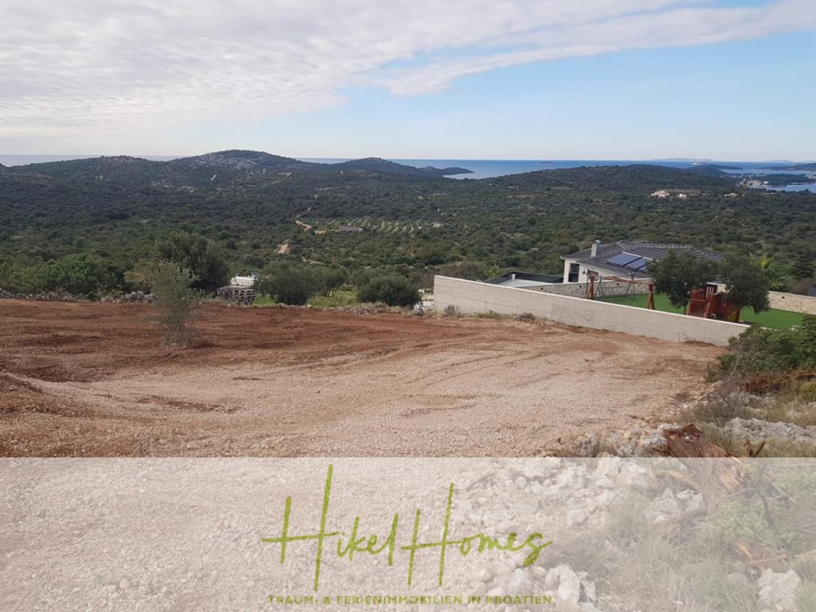 Prämiertes Architekturprojekt – Meerblick Luxus auf 490m² auf 1800m² Grund mit 2 Pools, Bar, Lounge - Ein gerodetes, felsiges Grundstück mit Blick auf grüne Hügel, verstreute Häuser und ein weit entferntes blaues Meer unter einem teilweise bewölkten Himmel. Auf der rechten Seite befindet sich ein modernes Haus mit einem Pool. Der Text am unteren Rand lautet "Hikel Homes". (45074)