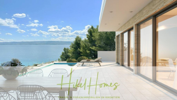 Eine moderne Terrasse mit weißen Möbeln bietet einen Blick auf den Pool, hohe Bäume und das blaue Meer unter einem sonnigen Himmel. Große Glastüren verbinden die Terrasse mit einem hellen Innenraum. Der Text lautet "Hikel Homes - Ferienimmobilien in Kroatien. - Schlüsselfertig Villa Annea – Meerblick von jedem Zimmer aus auf 250m² mit 3 Schlafzimmer / 3 Bädern