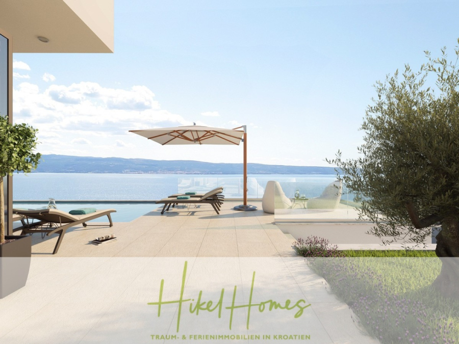 Schlüsselfertig Villa Annea – Meerblick von jedem Zimmer aus auf 250m² mit 3 Schlafzimmer / 3 Bädern - Eine moderne Terrasse am Meer mit Liegestühlen, einem Sonnenschirm und einem Infinity-Pool bietet einen Blick auf das ruhige blaue Wasser und die fernen Hügel. Ein Teil eines Gebäudes und ein Olivenbaum sind zu sehen. Der Text am unteren Rand lautet "Hikel Homes" in Grün. (46405)