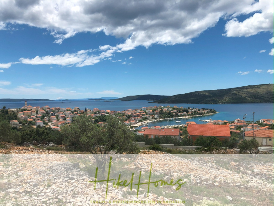 Meerblick – Schlüsselfertige Villa Princeza in Seget Vranijca – modernste Technik – 3-4 SZ - Blick auf eine Küstenstadt mit Terrakotta-Dächern, eingebettet am Meer und umgeben von Hügeln unter einem teilweise bewölkten Himmel. Im Vordergrund ist felsiges Gelände zu sehen und unten das Logo von "Hikel Homes". (45516)