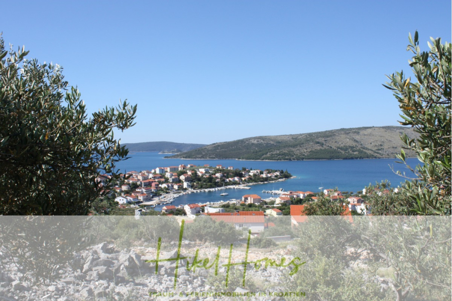 Meerblick – Schlüsselfertige Villa Princeza in Seget Vranijca – modernste Technik – 3-4 SZ - Eine Küstenstadt mit roten Dächern liegt an einer ruhigen blauen Bucht, umgeben von grünen Hügeln und Olivenbäumen unter einem klaren blauen Himmel. Im Vordergrund sieht man felsiges Gelände und belaubte Äste, die die malerische Aussicht einrahmen. (45521)