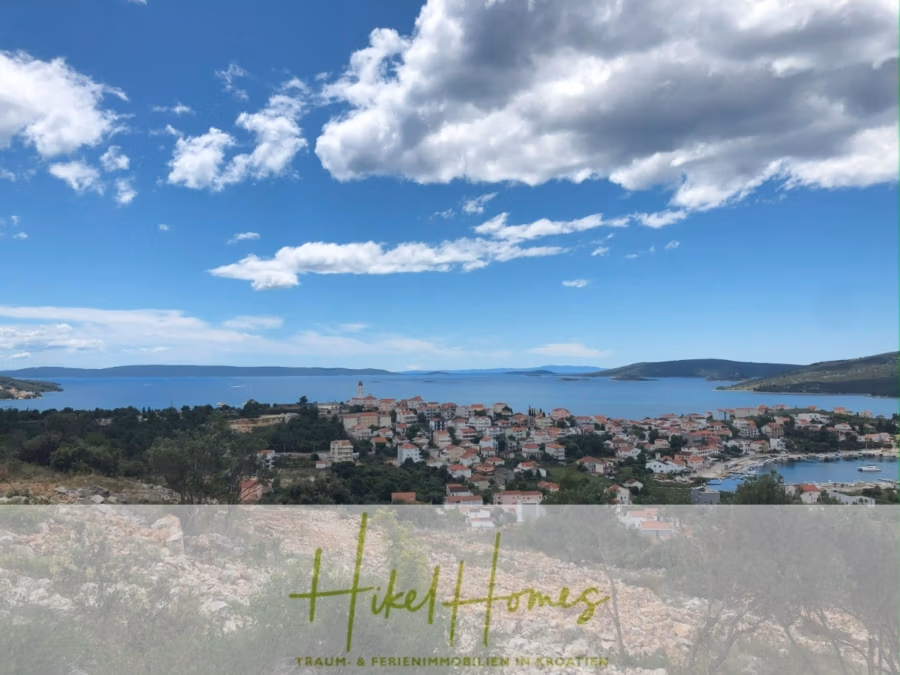 Meerblick – Schlüsselfertige Villa Princeza in Seget Vranijca – modernste Technik – 3-4 SZ - Malerische Aussicht auf eine Küstenstadt mit rotgedeckten Gebäuden am Meer. Die Landschaft ist von üppigem Grün und einem klaren, blauen Himmel mit vereinzelten Wolken geprägt. Im Hintergrund sind Hügel zu sehen. Unten steht „Hikel Homes“. (41325)