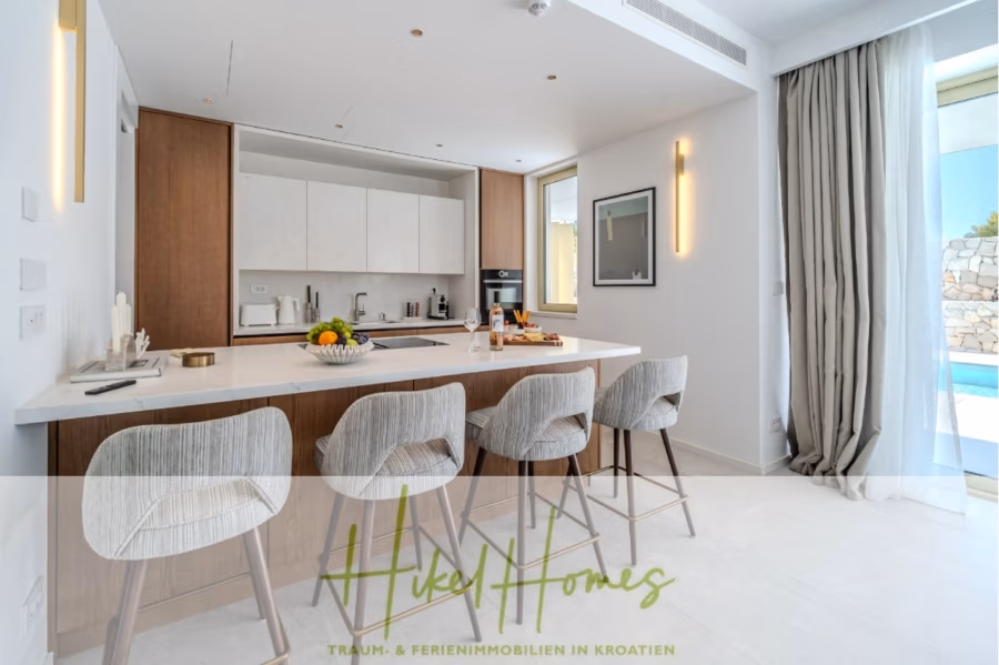 Schlüsselfertige Meerblick Villa SEA in Sutivan (Insel Brac) – 3 SZ + Bad, Pool, Sommerküche, … - Moderne Küche mit großer Kochinsel, vier grauen Barhockern und eleganten Schränken. Die Kochinsel verfügt über eine Obstschale und Küchenutensilien. Ein Fenster gibt den Blick auf eine Steinmauer und den Pool frei. Am Fenster hängt ein Vorhang, der Boden ist mit weißen Fliesen ausgelegt. (42999)
