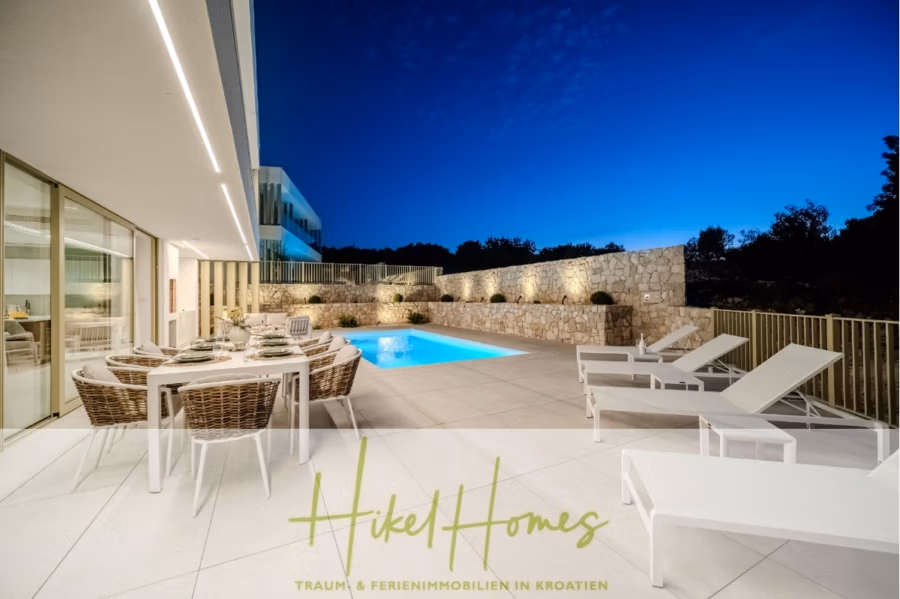 Schlüsselfertige Meerblick Villa SEA in Sutivan (Insel Brac) – 3 SZ + Bad, Pool, Sommerküche, … - Luxuriöse Außenterrasse mit Korbmöbeln und Liegestühlen neben einem wunderschön beleuchteten Swimmingpool. Moderne Architektur mit großen Fenstern, umgeben von einer Steinmauer. Abendhimmel im Hintergrund. Textüberlagerung: „Hikel Homes – Traum- & Ferienimmobilien in Kroatien.“. (42992)