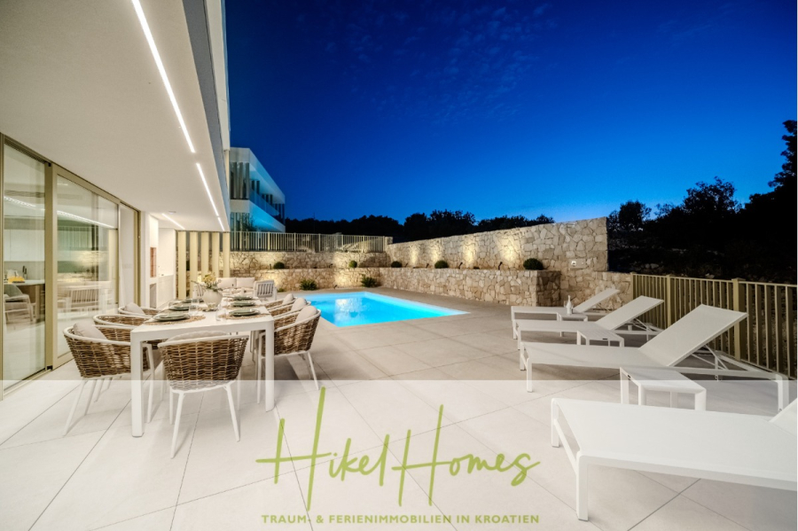 Schlüsselfertige Meerblick Villa SEA in Sutivan (Insel Brac) – 3 SZ + Bad, Pool, Sommerküche, … - Moderne Außenterrasse in der Abenddämmerung mit Esstisch, Korbstühlen und weißen Liegestühlen neben einem beleuchteten Swimmingpool; Glastüren führen in einen stilvollen Innenraum. Der Text am unteren Rand lautet: "Hikel Homes - Traum & Ferienimmobilien in Kroatien". (45017)
