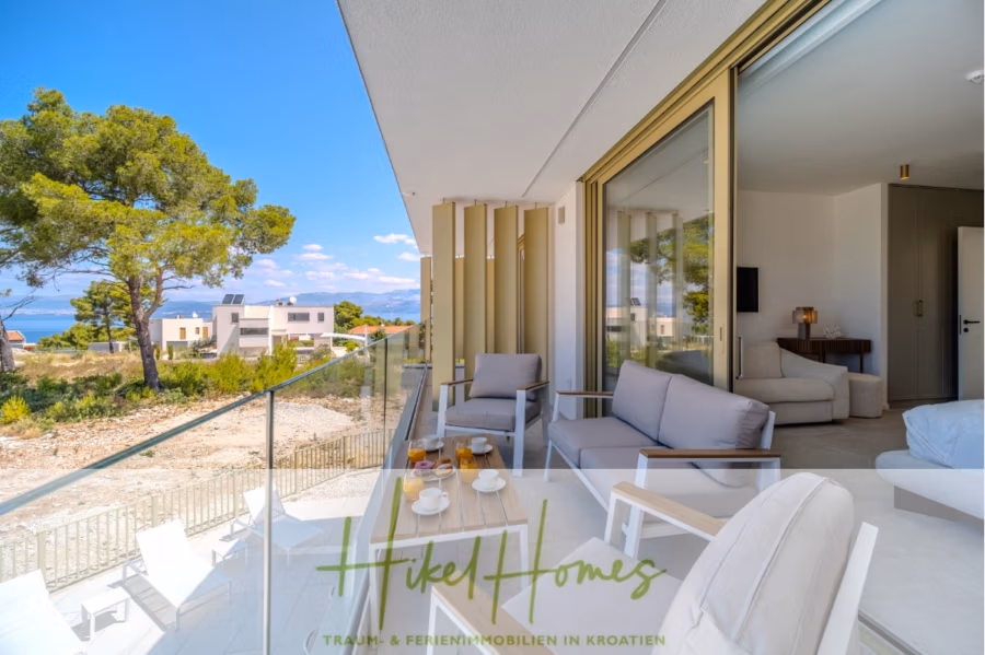 Schlüsselfertige Meerblick Villa SEA in Sutivan (Insel Brac) – 3 SZ + Bad, Pool, Sommerküche, … - Moderner Balkon mit Sitzgelegenheiten im Freien, Meerblick und umliegende Häuser. Blauer Himmel und Grün im Hintergrund. Glasgeländer und Schiebetüren öffnen sich zu einem hellen Raum. Teeservice und Obst auf dem Tisch. (43005)