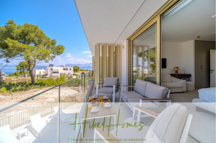 Schlüsselfertige Meerblick Villa SEA in Sutivan (Insel Brac) – 3 SZ + Bad, Pool, Sommerküche, … - Moderner Balkon mit Sitzgelegenheiten im Freien, Couchtisch für das Frühstück und Glasgeländer mit Blick auf Bäume, Häuser und das Meer unter blauem Himmel. Der innere Wohnbereich ist durch große Schiebetüren einsehbar. (45031)